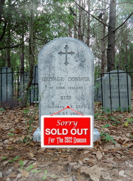 A weathered tombstone in a wooded cemetery with a 'Sold Out' sign in front.