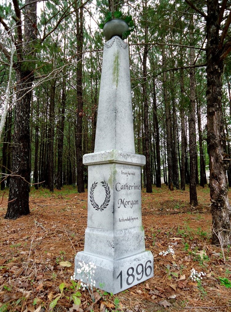 White obelisk monument in a forested area with engraved dates. White obelisk monument in a forested area with engraved dates.