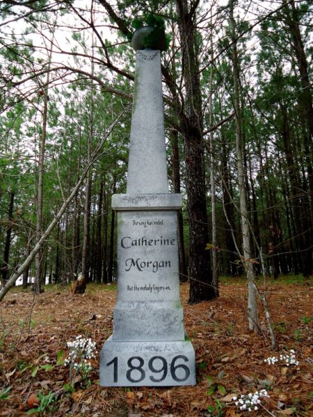 A tall white monument in a wooded area dated 1896.