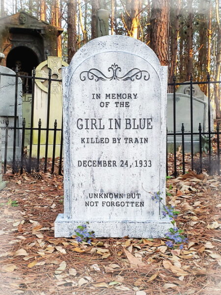 A decorative tombstone in a forest setting with a wrought iron fence.