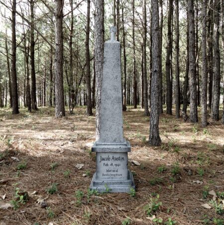A weathered gravestone stands among tall pine trees in a quiet forest.