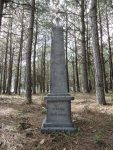 A weathered gravestone stands among tall pine trees in a quiet forest.