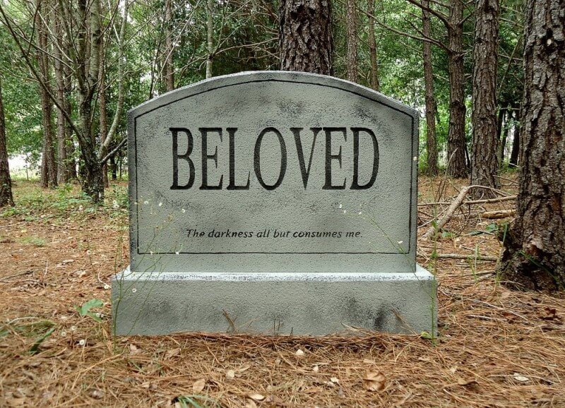 A gray tombstone with the word 'BELOVED' in a forested cemetery. A gray tombstone with the word 'BELOVED' in a forested cemetery.